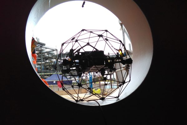 A cage-protected inspection drone hovering at a circular man hatch, seen from inside the structure